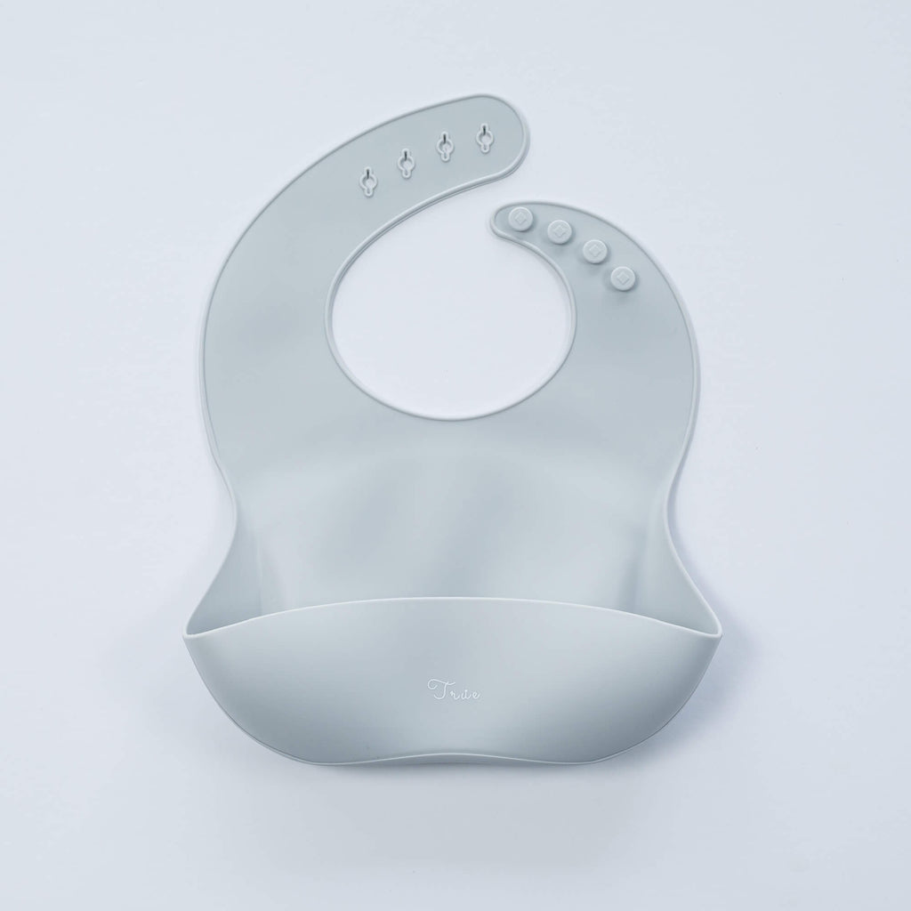 A light gray, BPA-free Silicone Bucket Bib with an adjustable neck strap featuring snap buttons. The bib has a scoop at the bottom to catch food.