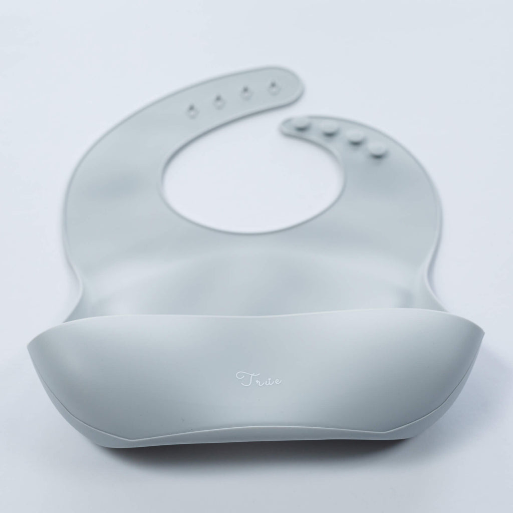 A light gray, BPA-free Silicone Bucket Bib with an adjustable neck strap featuring snap buttons. The bib has a scoop at the bottom to catch food.