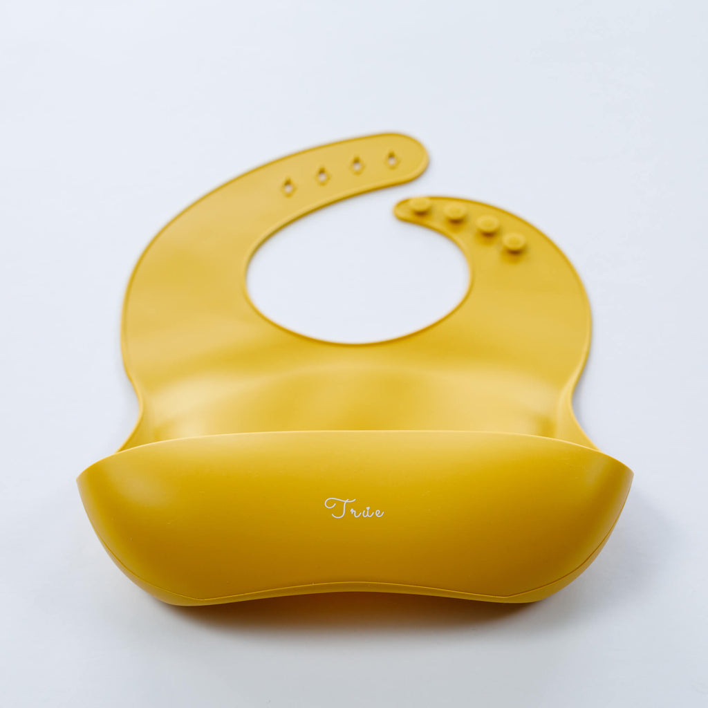 A yellow, BPA-free Silicone Bucket Bib with an adjustable neck strap featuring snap buttons. The bib has a scoop at the bottom to catch food.