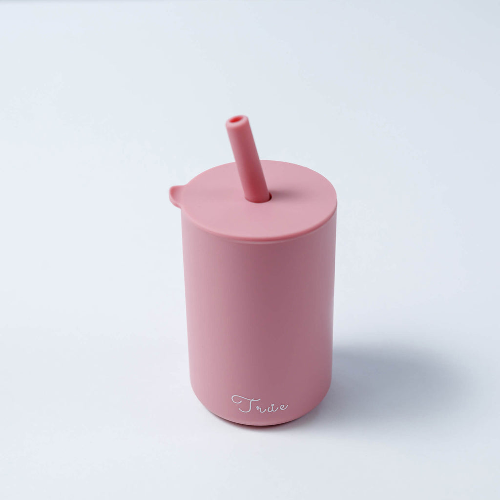 A cylindrical pastel pink cup with a straw and a lid, featuring the word ‘True’ in a light font near the bottom.