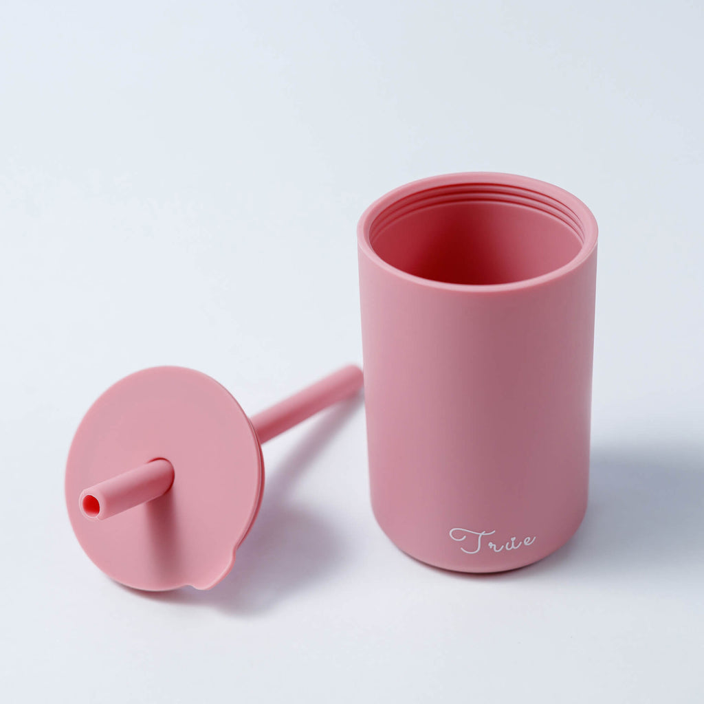 A cylindrical pastel pink cup with a straw and a lid, featuring the word ‘True’ in a light font near the bottom.