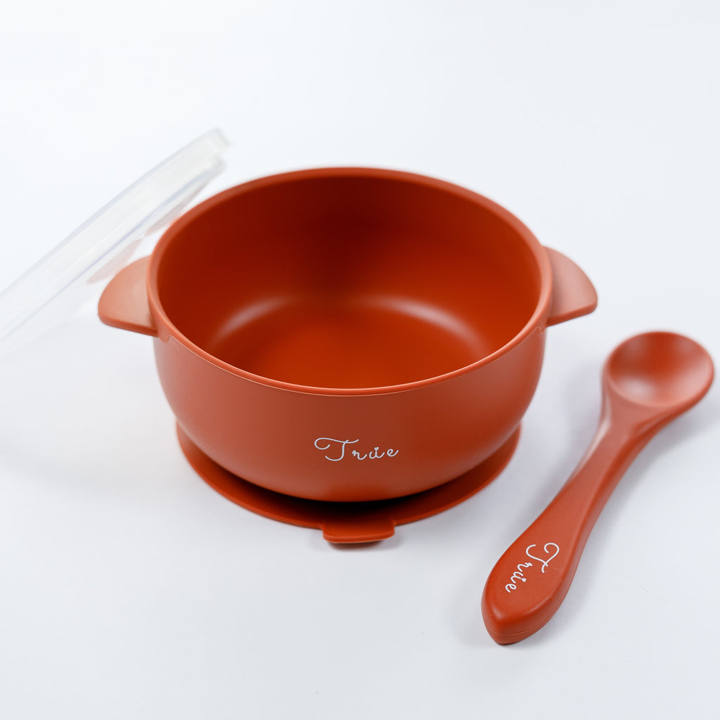 A red silicone suction bowl with two handles and a lid, including a spoon labeled with the brand ‘True'.