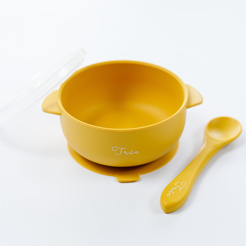 A yellow silicone suction bowl with two handles and a lid, including a spoon labeled with the brand ‘True'.