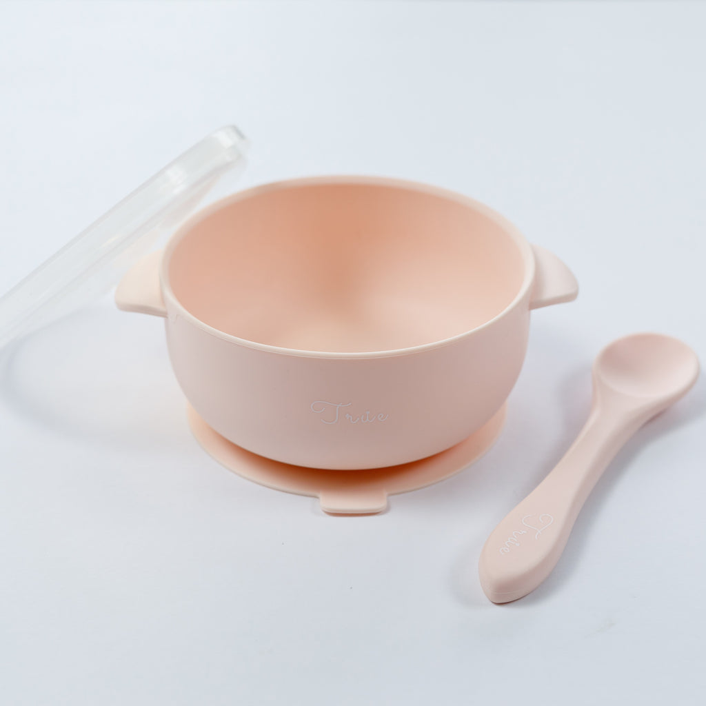 A pastel pink silicone suction bowl with two handles and a lid, including a spoon labeled with the brand ‘True'.