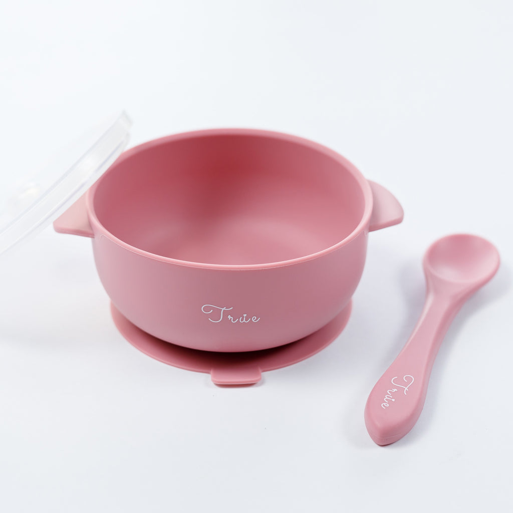 A pink silicone suction bowl with two handles and a lid, including a spoon labeled with the brand ‘True'.