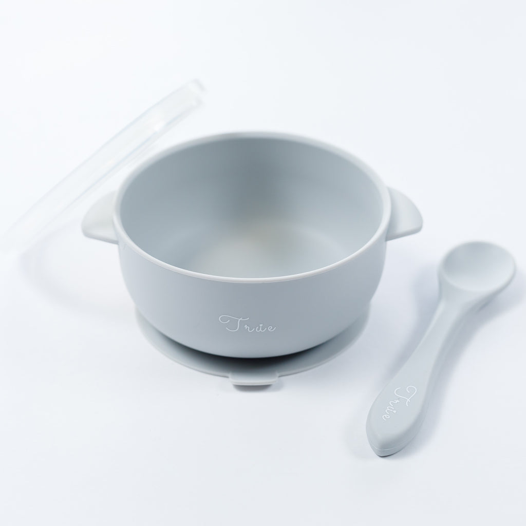 A light grey silicone suction bowl with two handles and a lid, including a spoon labeled with the brand ‘True'.