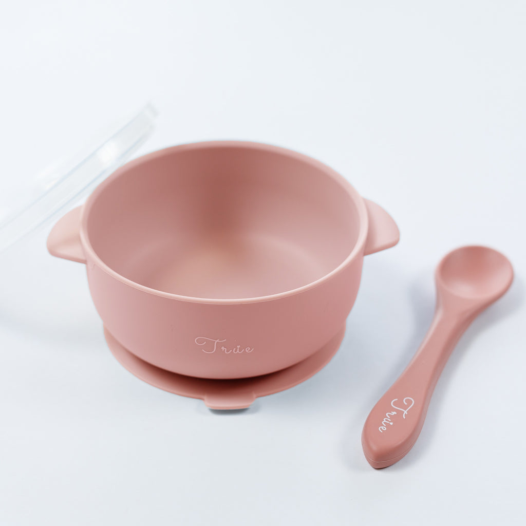 A pink silicone suction bowl with two handles and a lid, including a spoon labeled with the brand ‘True'.