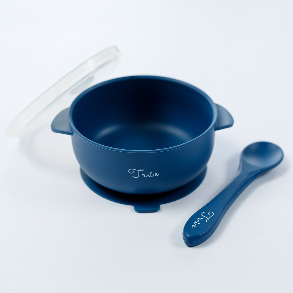 A midnight blue silicone suction bowl with two handles and a lid, including a spoon labeled with the brand ‘True'.