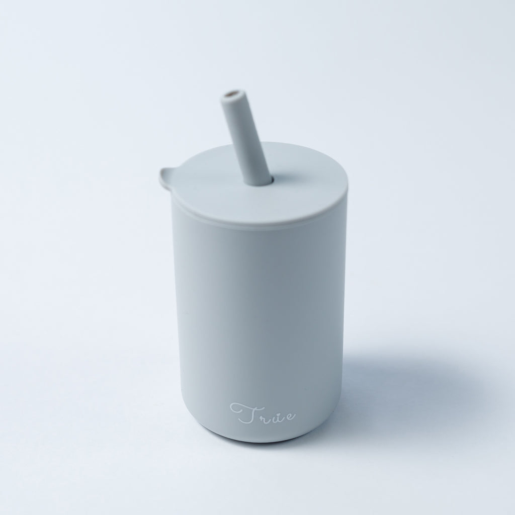 A cylindrical light grey cup with a straw and a lid, featuring the word ‘True’ in a light font near the bottom.