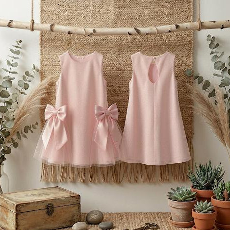 Two pink dresses with bows hanging on a textured wall with plants and a wooden box below.