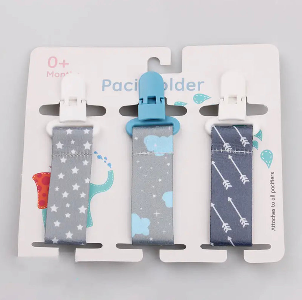 Three pacifier clips with varying patterns, attached to a cardboard holder with a blue and white design featuring a unicorn and stars.