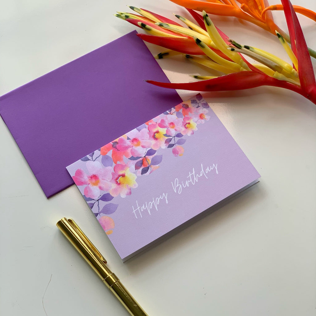 Purple birthday card with floral design and 'Happy Birthday' text, next to a gold pen and colorful flowers on a white background.