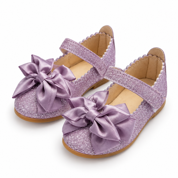 A pair of toddler girl's glittery lavender Mary Jane shoes with large satin bow embellishments on the toes, shown on a clean white background.