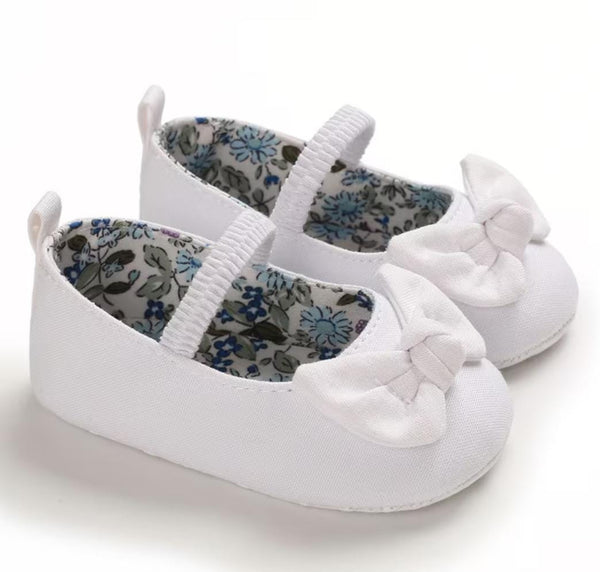 A pair of white baby ballet flats with elastic straps and large white fabric bows on the toes, shown on a clean white background.