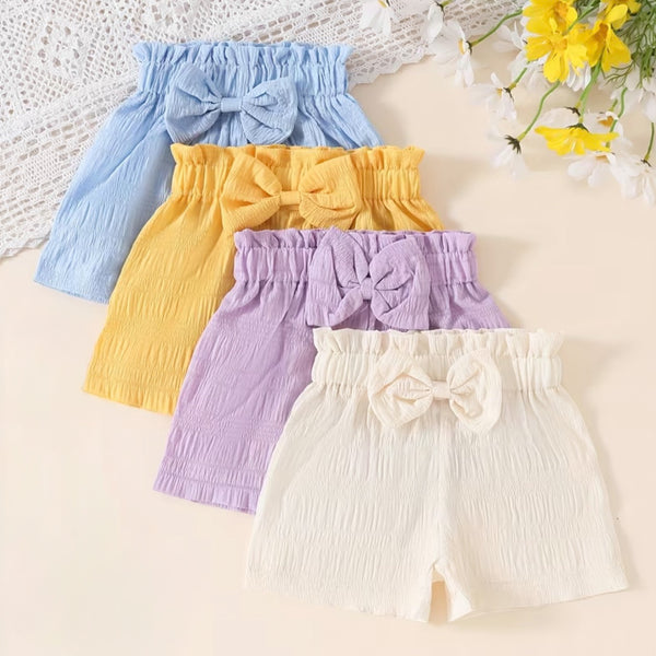 Four pairs of baby shorts in blue, yellow, purple, and white with bow details on a light background.
