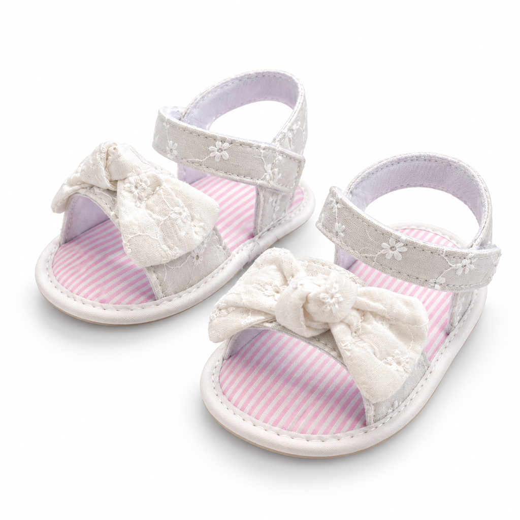 A pair of baby sandals with white fabric straps, white embroidered floral details, and large white lace bows on the toes. The soles have a white and pink striped pattern, and the inside of the straps is light purple.