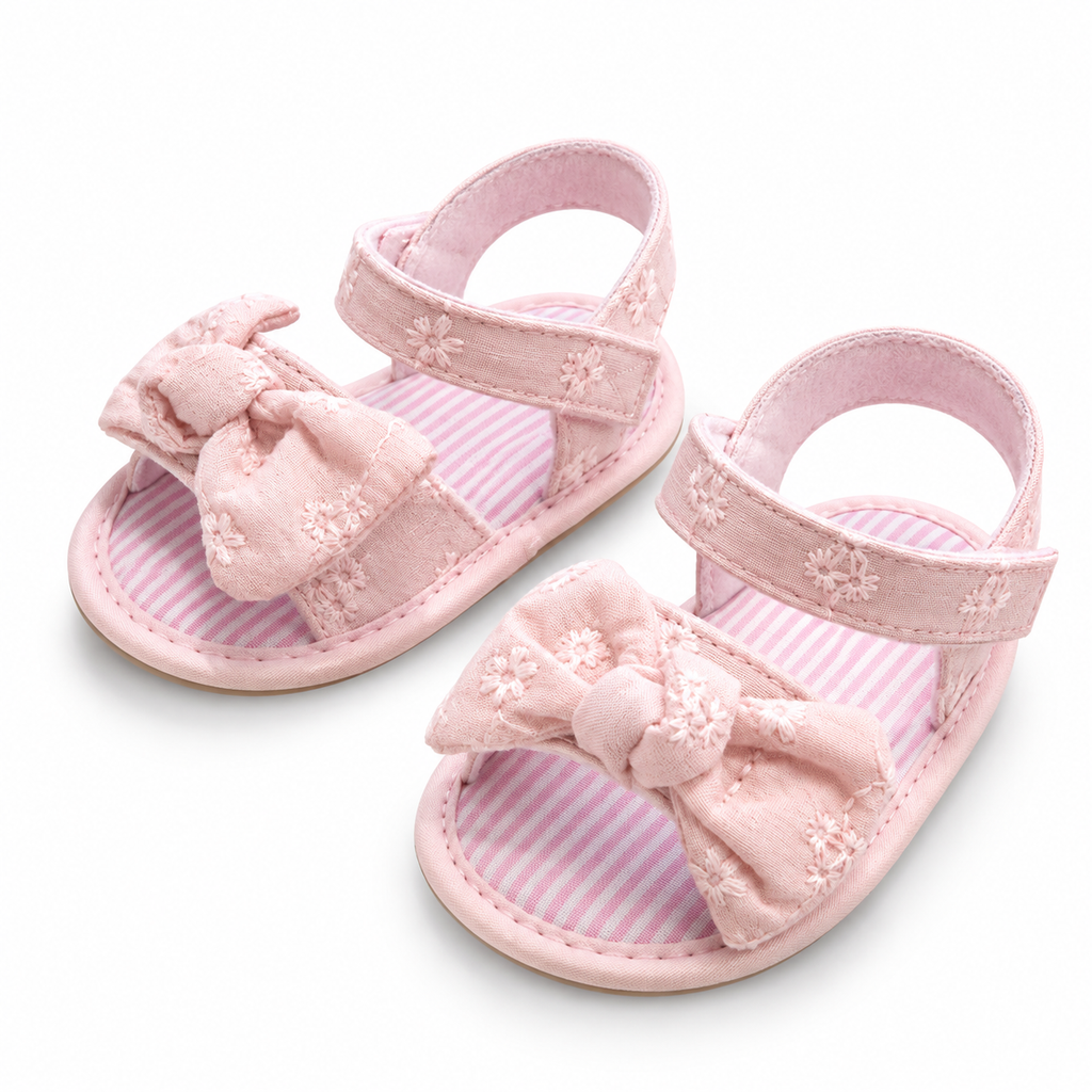 A pair of baby sandal shoes in light pink, featuring a large fabric bow on the toe strap, delicate embroidered floral details, and a pink and white striped footbed.