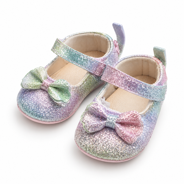 A pair of toddler glitter Mary Jane shoes with iridescent pastel rainbow finishes, featuring wide straps and decorative bows on the toes, shown on a white background.