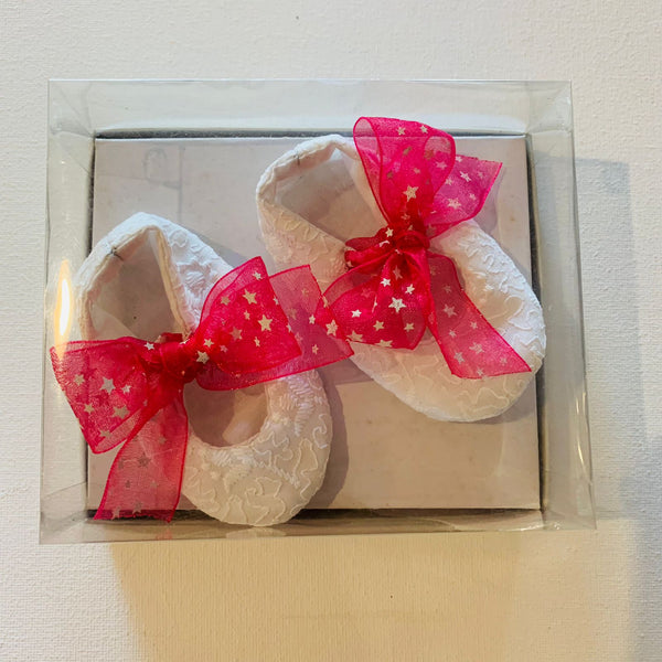 A pair of handmade white baby shoes with a pink bow and red star appliques, presented in a clear plastic box.