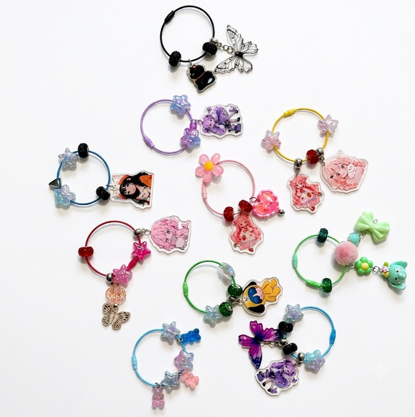 A collection of colorful hair claw clips arranged on a plain white background. The clips feature various charms including butterflies, flowers, hearts, cartoon characters, and pom-poms in a wide range of colors.