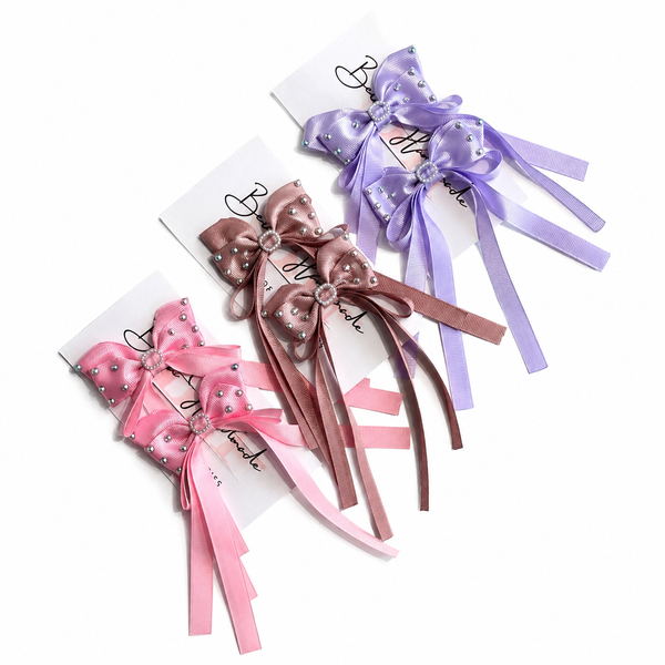 Three satin ribbon hair bows in pink, dusty rose, and lavender purple, each decorated with small rhinestones and attached to a white card on a plain white background.