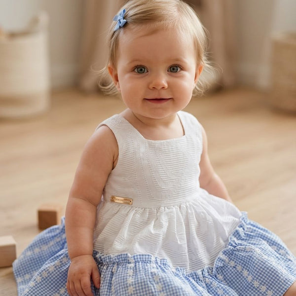 Baby in a white and blue dress sitting on a wooden floor.