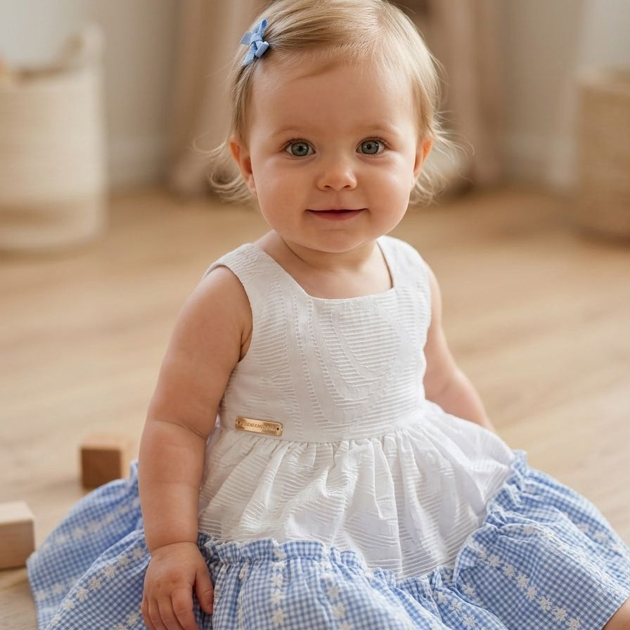 Baby in a white and blue dress sitting on a wooden floor.