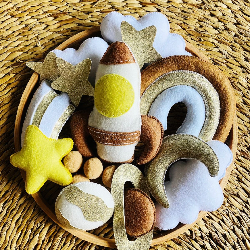 A handcrafted felt mobile with a space theme, including a rocket, stars, moons, and clouds, displayed on a wicker surface.