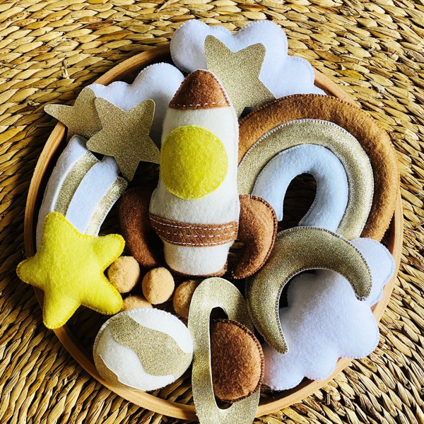 A handcrafted felt mobile with a space theme, including a rocket, stars, moons, and clouds, displayed on a wicker surface.