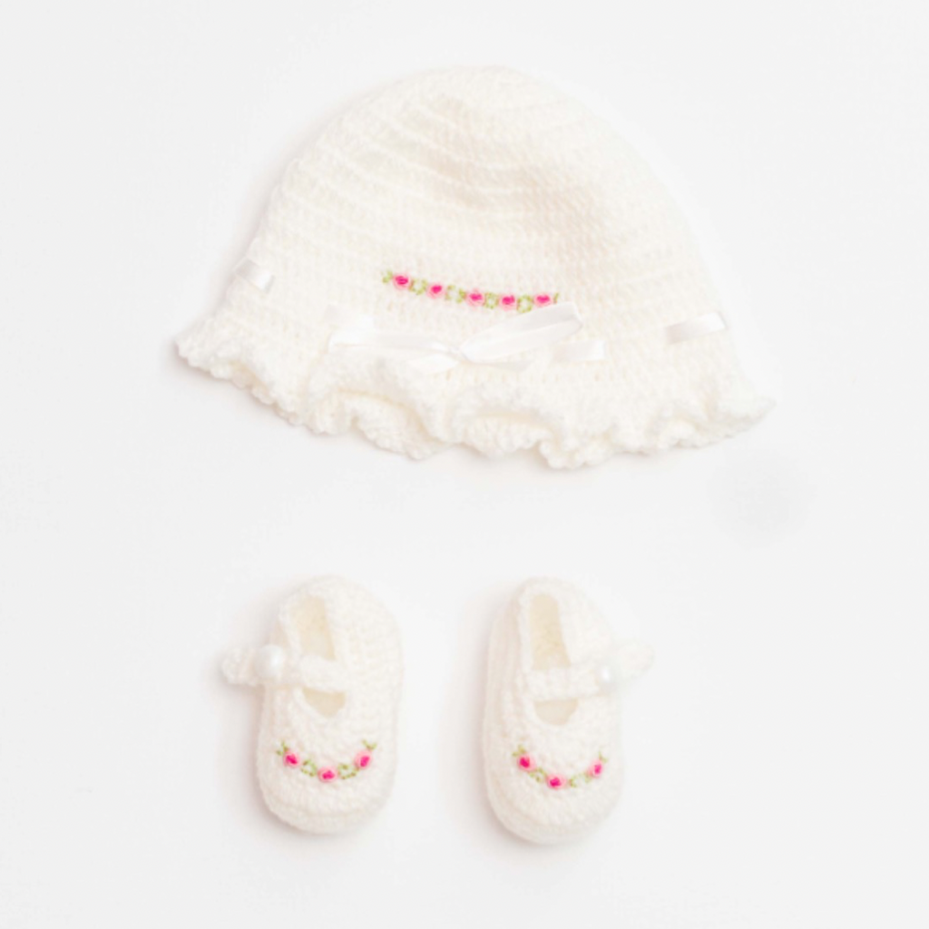 A white crochet embroidered cap and booties set for newborns with pink details.