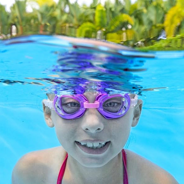 A child wearing purple and pink swim goggles underwater in a bright blue swimming pool, with palm trees visible in the background.