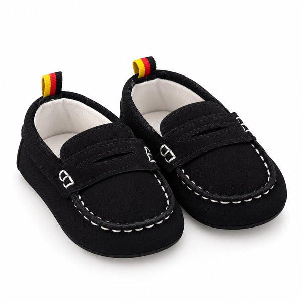 A pair of black baby moccasin shoes with white contrast stitching, silver buckle details, and a small yellow, red, and black pull tab on the heel, shown on a white background.