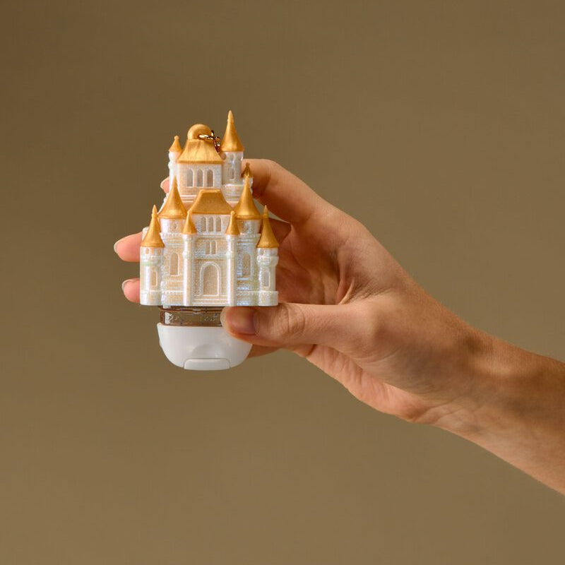 A hand holding a small white and gold official disney castle-shaped PocketBac Holder Keyring against a solid warm brown background.