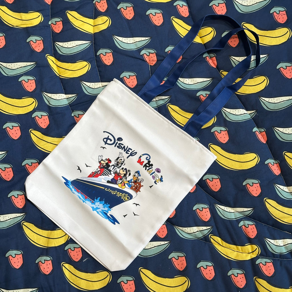 A white canvas tote bag with a Disney Perseus graphic print, laid flat on a navy blue fabric background patterned with yellow bananas and red strawberries.
