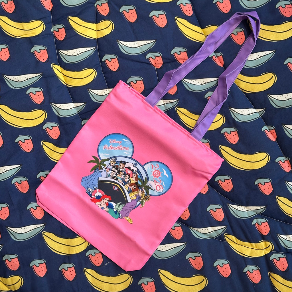 A bright pink tote bag with purple handles, featuring a colorful Mickey Mouse-themed graphic, laid flat on a navy blue quilted blanket patterned with bananas, strawberries, and ice cream cones.
