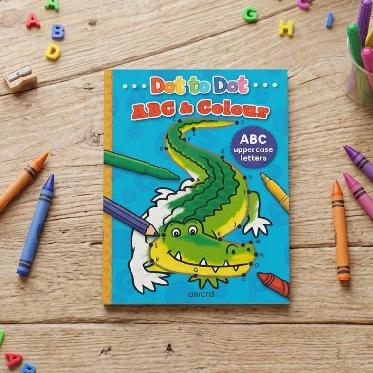 A children's coloring book titled 'Dot to Dot ABC & Colours' by Oryx & Cavo lies on a light wooden tabletop, surrounded by colorful crayons and alphabet letters. The cover features a bright blue background with a cartoon green alligator being colored in with crayons.