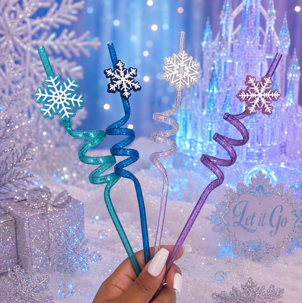 A hand holds four glittery winter-themed party straws with snowflake tops, set against a festive Frozen-themed backdrop with a castle and snow decorations.