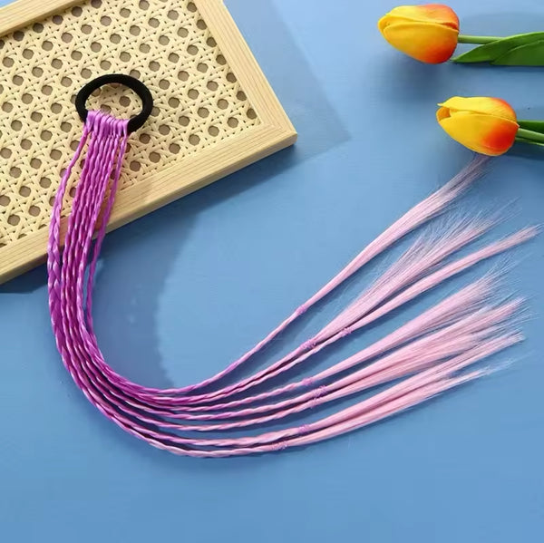 The Hair Braid Extension Band - Ombre Pink & Purple by True rests on a beige woven mat against a blue surface, featuring multiple lengthy, braided strands. Two yellow and orange tulips are placed to the right of the hair accessory, making it perfect for kids' hairstyles.