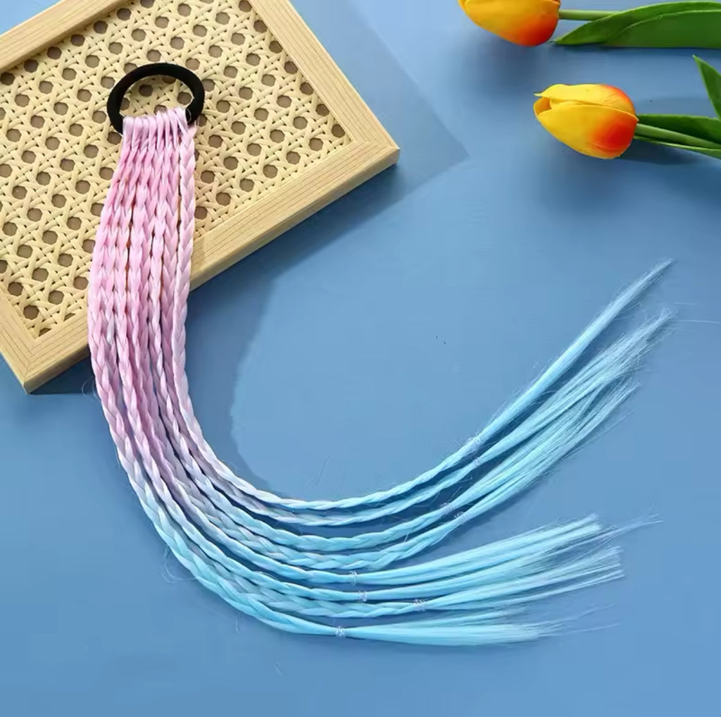 The True Hair Braid Extension Band - Ombre Pink & Blue features a stylish braid transitioning from pink to blue, attached to a black hair tie. It rests on a square frame with a woven pattern, set against a blue surface. In the corner, there is a small bunch of yellow tulips with green leaves—perfect for adding flair to kids' hair extensions.