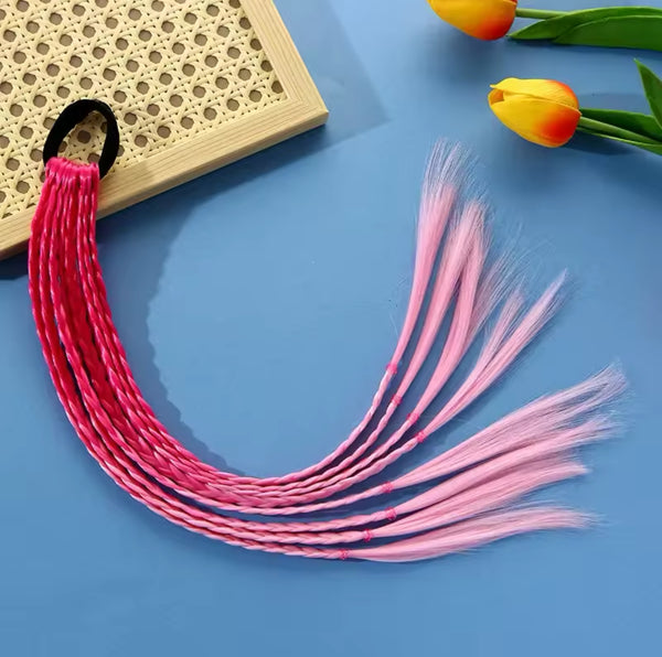 A bundle of True's Hair Braid Extension Band in Ombre Pink, secured with a black elastic band, is displayed on a blue surface. The extensions are fanned out with some strands braided and others left loose, ideal for creating a stylish braid design. Yellow and orange tulips add a vibrant touch in the upper right corner.