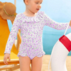 A toddler girl wearing a long sleeve purple and white floral rashguard swimsuit with frill bottoms, 