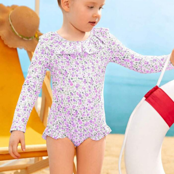 A toddler girl wearing a long sleeve purple and white floral rashguard swimsuit with frill bottoms, 