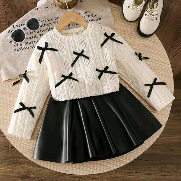 White sweater with black cross details and black pleated skirt on a wooden surface.