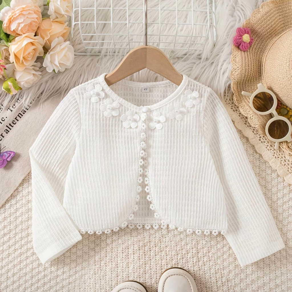 White baby cardigan on a hanger with flowers and a hat in the background