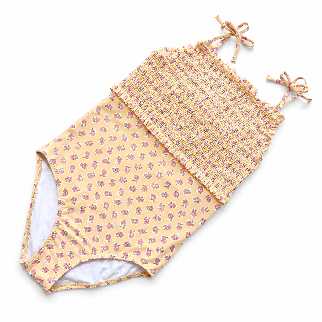 A one-piece baby swimsuit with a smocked bodice and tie straps, featuring a yellow and pink floral print on a white background.