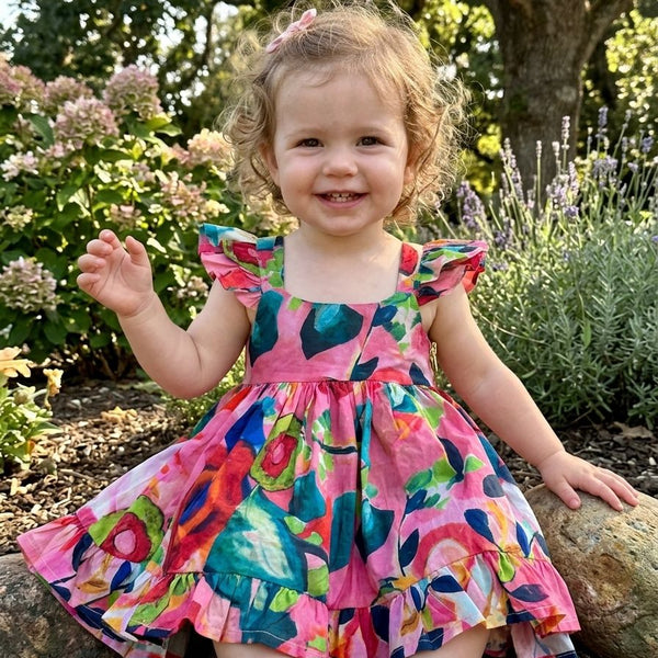 Child in a colorful dress sitting outdoors with flowers and trees in the background