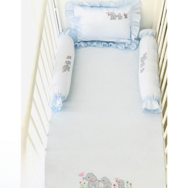 The Elephant Family Cotton Bedding Set - Blue by True transforms a baby crib into a cozy and charming haven. This set includes a pale blue sheet, pillow with ruffled edges, and two side bolsters, all made from soft cotton and adorned with cute elephants holding pink flags. The crib, surrounded by white bars, becomes an adorable sanctuary for your little one.