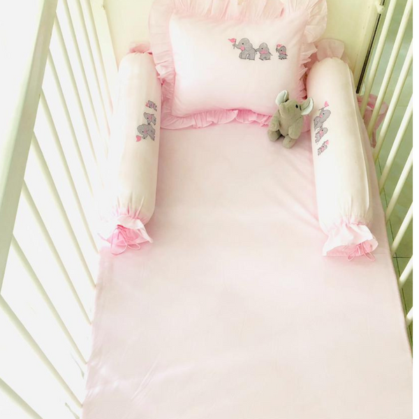 The True Elephant Family Cotton Bedding Set in pink adorns a baby's bed, complete with light pink sheets, pillows, and bolsters featuring adorable elephant embroidery. A stuffed elephant toy sits on the bed, perfectly matching the nursery's elephant theme. The crib has white rails and a cozy cotton bedding set, creating a soft and comfortable sleeping area for your baby.