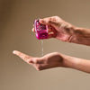 A person's hands holding and dispensing a pink hand sanitizer bottle against a solid beige background.
