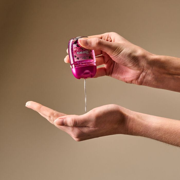 A person's hands holding and dispensing a pink hand sanitizer bottle against a solid beige background.